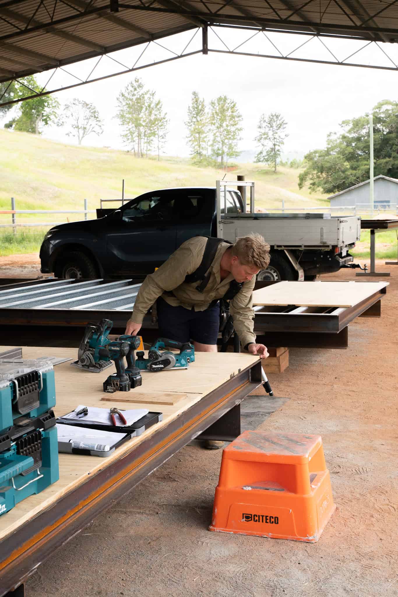 A carpenter marking out on a module floor deck under the shed roof, ute and paddock behind